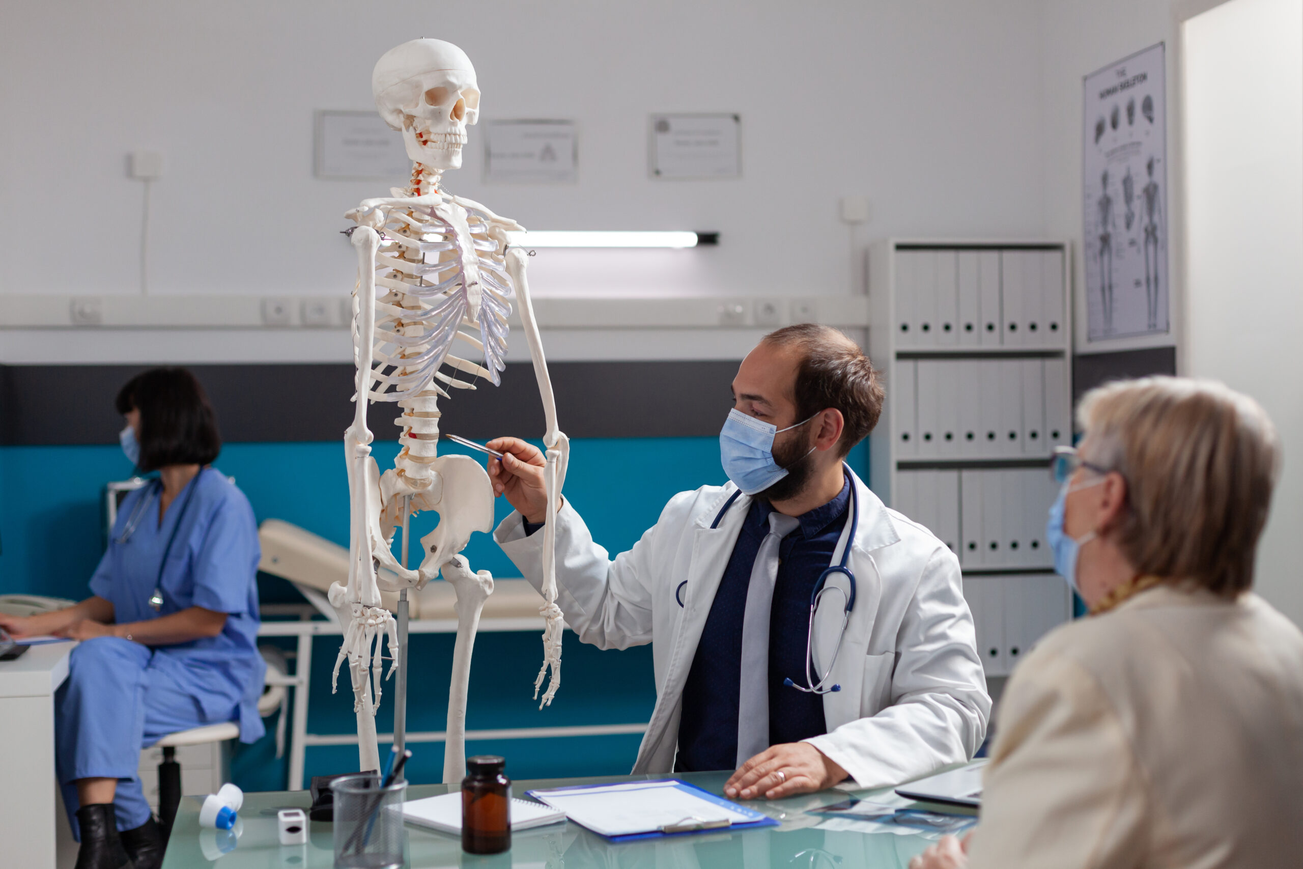 Specialist explaining human skeleton to retired woman at checkup visit, talking about bones injury at osteopathy appointment during covid 19 pandemic. Doctor showing joint model to patient.